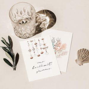 May include: A white card with the text "our brilliant summer" and a watercolor illustration of seashells hanging from a string. There is a glass of water, a green sprig, and a seashell on a white background.