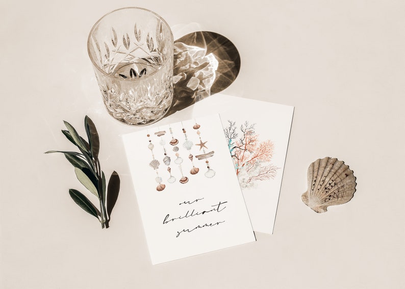 May include: A white card with the text "our brilliant summer" and a watercolor illustration of seashells hanging from a string. There is a glass of water, a green sprig, and a seashell on a white background.
