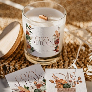 Peut inclure: Un bocal en verre contenant une bougie blanche avec une &eacute;tiquette indiquant "Cozy Autumn". L'&eacute;tiquette pr&eacute;sente une illustration aquarelle de citrouilles, de feuilles et de fleurs. La bougie est pos&eacute;e sur une surface en osier brune.