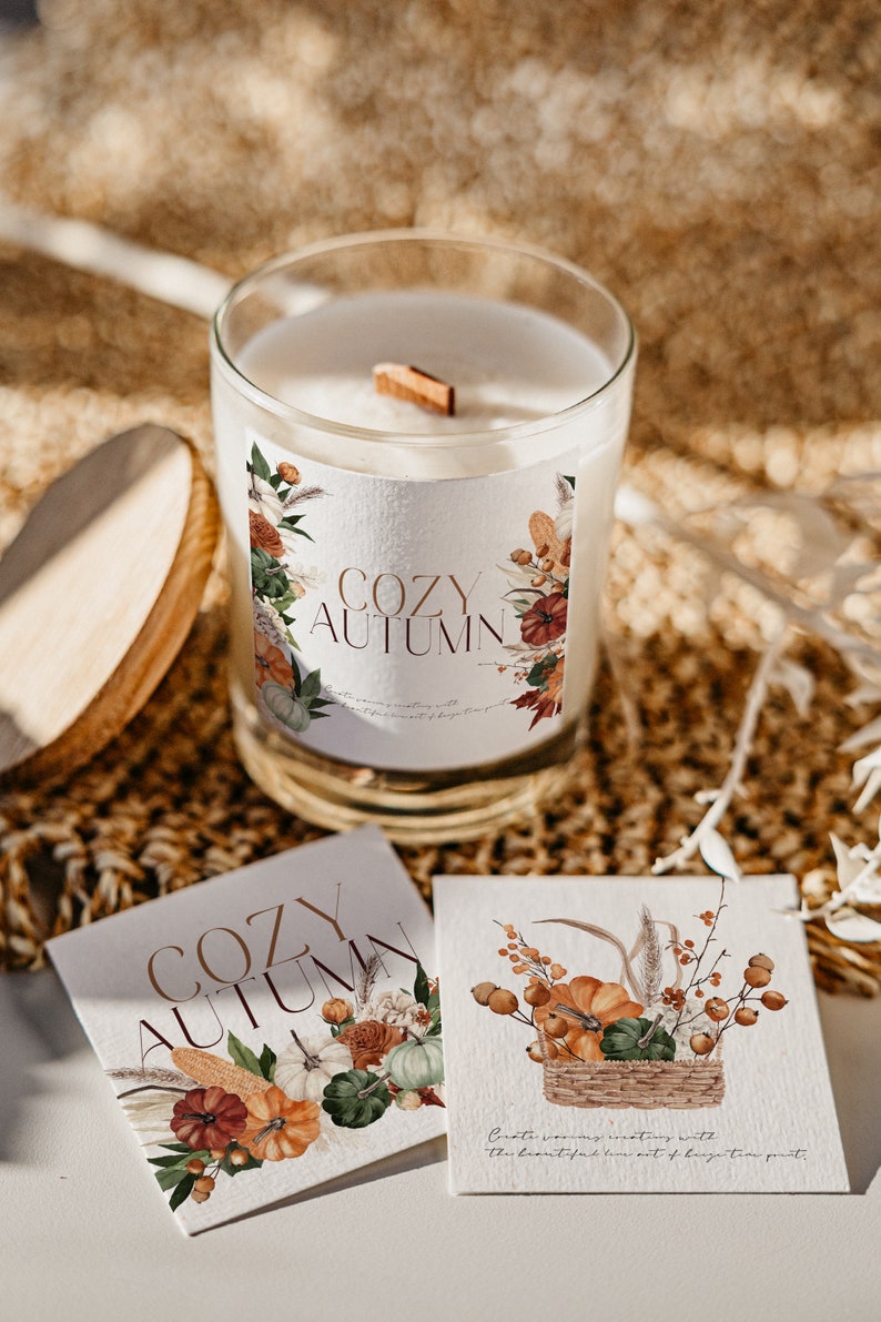 Peut inclure: Un bocal en verre contenant une bougie blanche avec une &eacute;tiquette indiquant "Cozy Autumn". L'&eacute;tiquette pr&eacute;sente une illustration aquarelle de citrouilles, de feuilles et de fleurs. La bougie est pos&eacute;e sur une surface en osier brune.