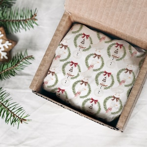 May include: A cardboard box with a Christmas-themed gift inside. The gift is wrapped in white paper with a repeating pattern of snowmen wearing wreaths and red bows. A gingerbread snowflake cookie and pine branches are in the background.