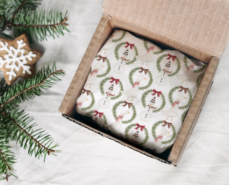 May include: A cardboard box with a Christmas-themed gift inside. The gift is wrapped in white paper with a repeating pattern of snowmen wearing wreaths and red bows. A gingerbread snowflake cookie and pine branches are in the background.