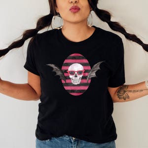 May include: Black t-shirt featuring a skull wearing red sunglasses and bat wings, set against a pink and black striped oval. The design is centered on the shirt. The model is wearing blue jeans.