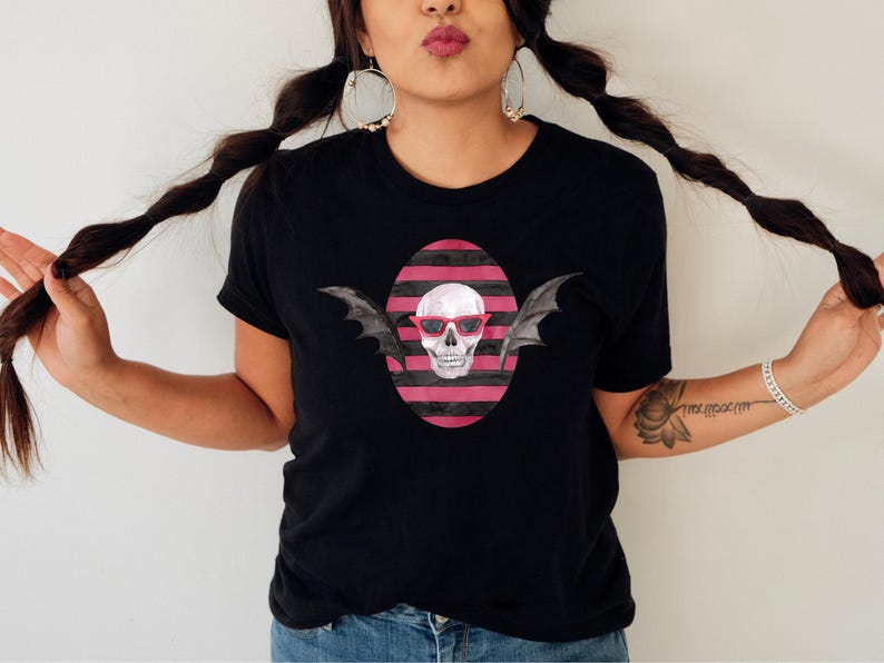 May include: Black t-shirt featuring a skull wearing red sunglasses and bat wings, set against a pink and black striped oval. The design is centered on the shirt. The model is wearing blue jeans.