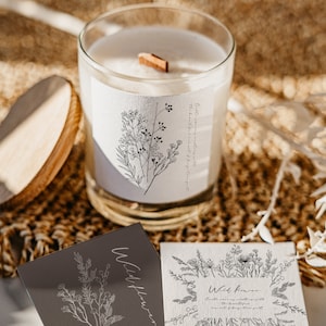 May include: A white candle in a glass jar with a wooden wick. The candle is labelled "Wildflower" and features a floral design. The candle is resting on a brown woven surface with two cards that say "Wildflower" on them.