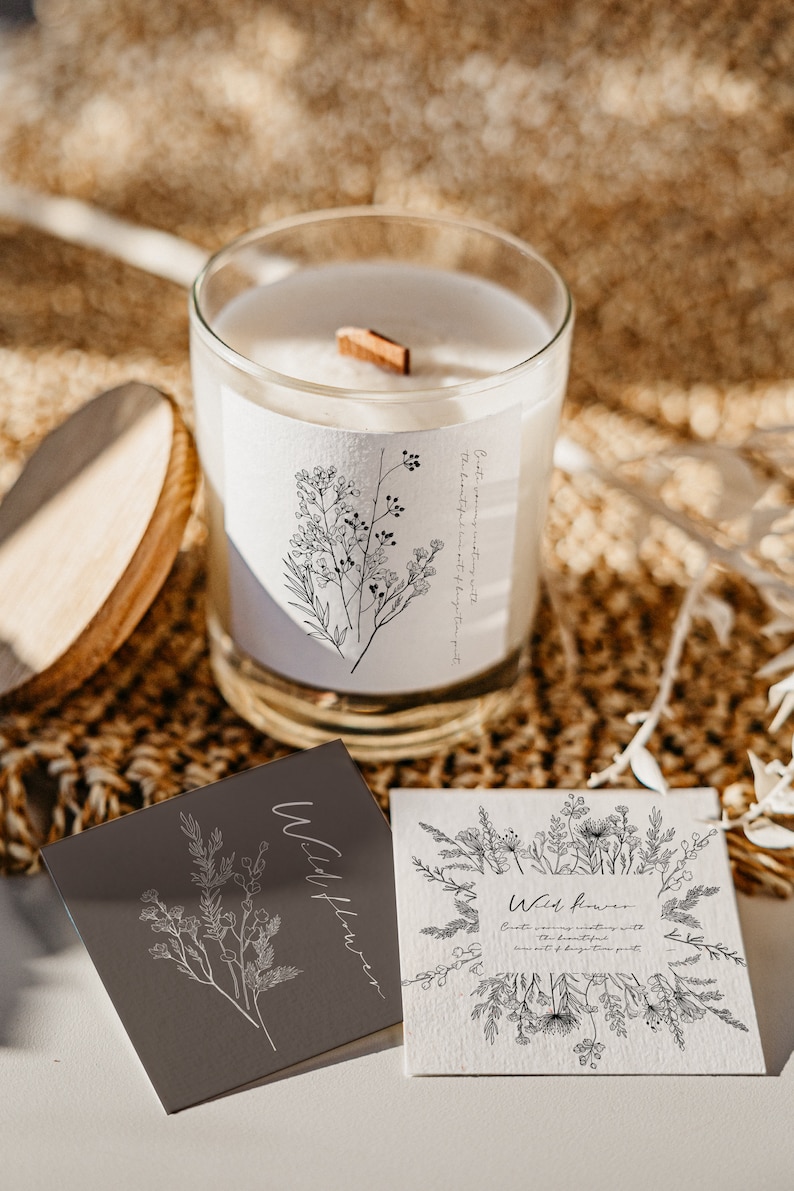 May include: A white candle in a glass jar with a wooden wick. The candle is labelled "Wildflower" and features a floral design. The candle is resting on a brown woven surface with two cards that say "Wildflower" on them.