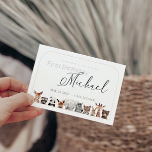 May include: A white invitation card with a light brown border, held by a hand. The card reads "First Birthday Michael" with the date May 23, 2023. A row of watercolor animals decorates the bottom.