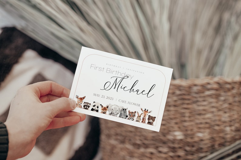 May include: A white invitation card with a light brown border, held by a hand. The card reads "First Birthday Michael" with the date May 23, 2023. A row of watercolor animals decorates the bottom.