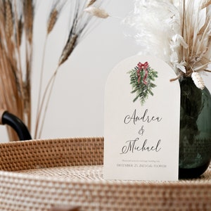 May include: A white wedding invitation with a watercolor illustration of a Christmas wreath with a red bow. The invitation reads "Andrea & Michael" and "December 23, 2023 Gala, Flower"