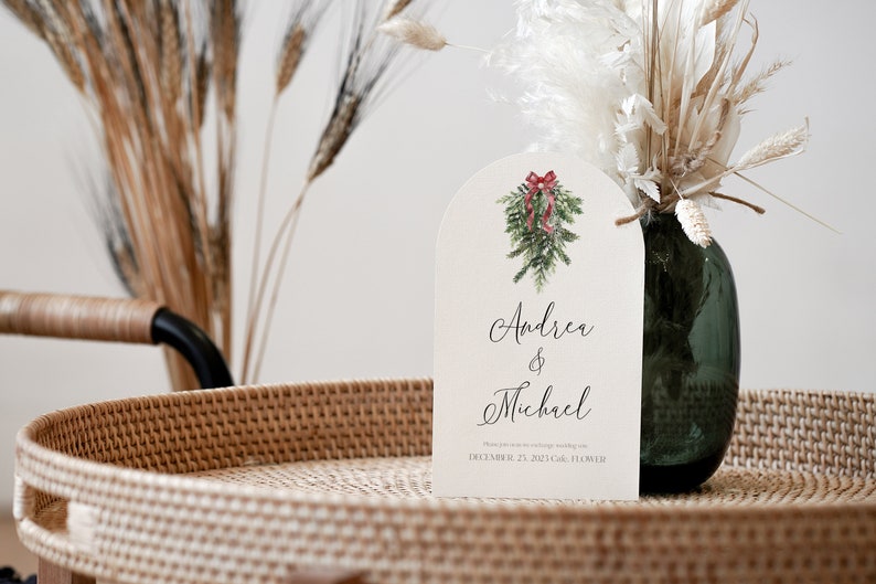 May include: A white wedding invitation with a watercolor illustration of a Christmas wreath with a red bow. The invitation reads "Andrea & Michael" and "December 23, 2023 Gala, Flower"