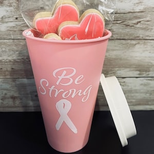 May include: Pink tumbler with a white lid and a white design that says "Be Strong" with a pink ribbon. The tumbler is filled with pink heart-shaped cookies.