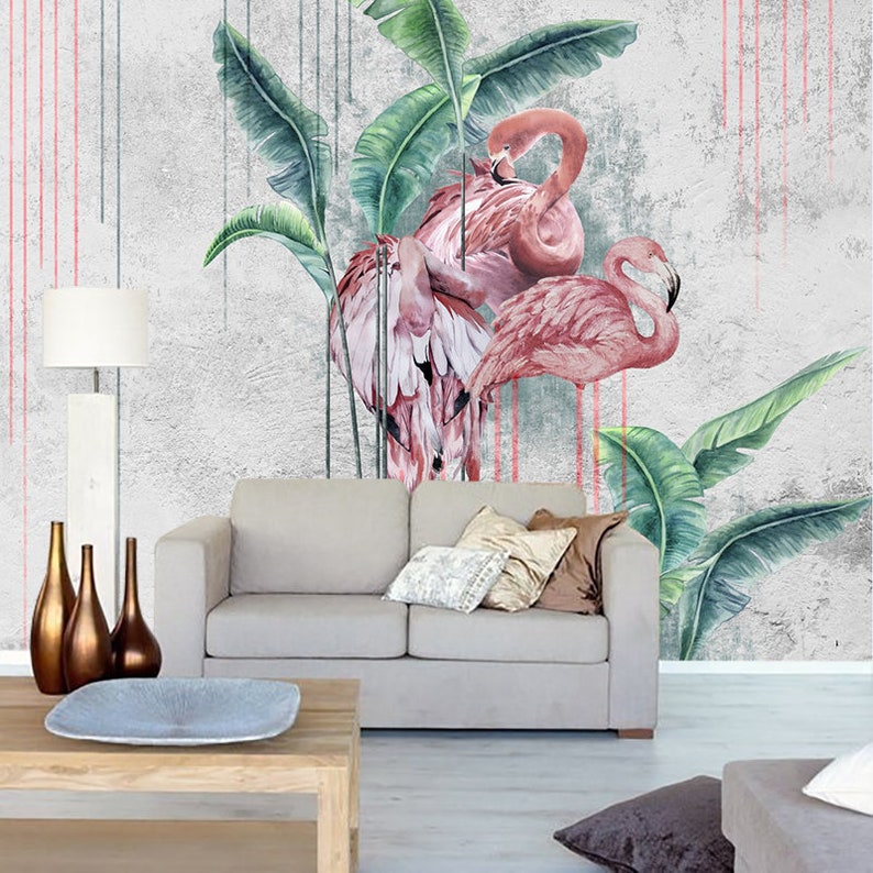 May include: A watercolor painting of two pink flamingos standing in front of a gray concrete wall with green banana leaves. The wall has red and pink paint streaks.