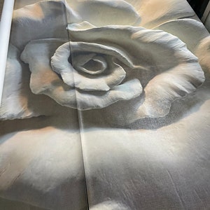 Op de afbeelding: Een close-up van een grote, witte, stoffen roos met een textuur. De roos is gedeeltelijk opgerold, waardoor de stof eronder zichtbaar wordt.