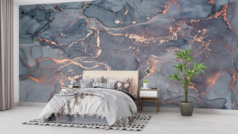 May include: A bedroom with a large, abstract marble wall mural in shades of gray and copper. The room features a bed with a light-colored wooden headboard, a nightstand, a lamp, and a potted plant. The bed has a gray and white striped blanket.