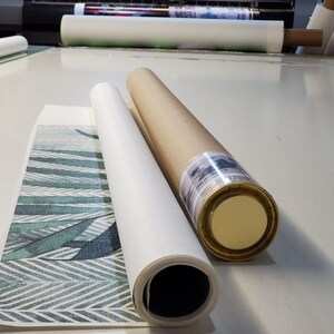 May include: Two rolled canvases, one with a green and white geometric pattern and the other with a brown paper covering, are on a white surface. The canvas with the pattern is partially unrolled, revealing the design.