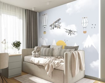 May include: A light blue and white nursery wall mural with a yellow sun, white clouds, and gray mountains. There are two gray airplanes and two hot air balloons flying in the sky. The mural is in a room with a white couch and a white teddy bear.