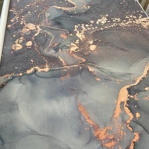 May include: A roll of decorative wallpaper featuring an abstract design. The pattern showcases flowing, smoky gray and blue hues with accents of copper-colored metallic detailing. The wallpaper is rolled up, ready for use.
