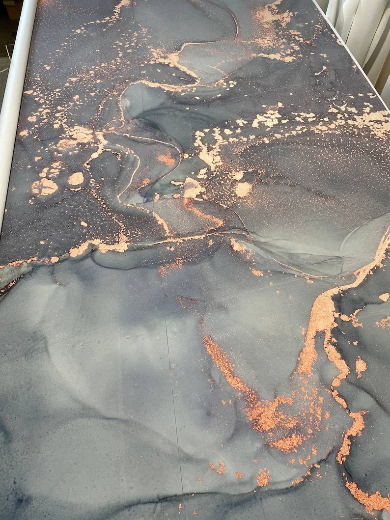 May include: A roll of decorative wallpaper featuring an abstract design. The pattern showcases flowing, smoky gray and blue hues with accents of copper-colored metallic detailing. The wallpaper is rolled up, ready for use.