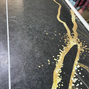 May include: A black fabric with a gold design of a winding river with scattered gold dots. The fabric is being rolled up on a white tube.