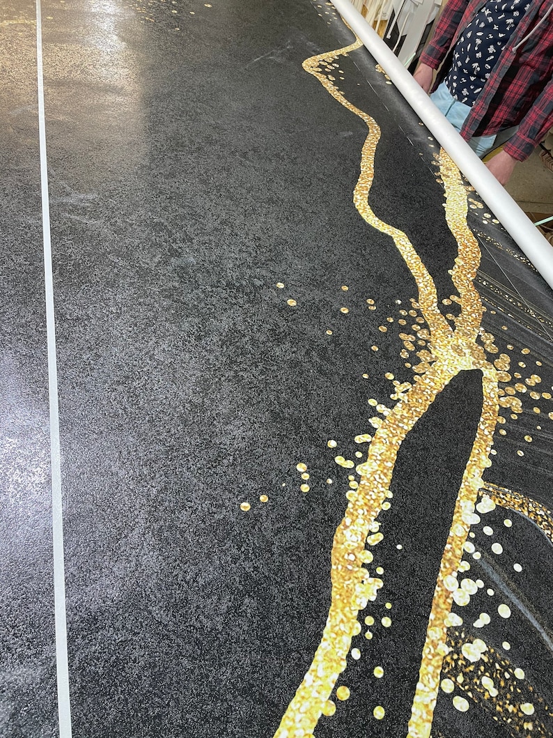 May include: A black fabric with a gold design of a winding river with scattered gold dots. The fabric is being rolled up on a white tube.