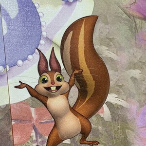 May include: A cartoon squirrel with brown fur and a bushy tail stands on its hind legs with its arms outstretched. The squirrel has a wide smile and large, round eyes. The background is a blurred image of a purple and white fabric.