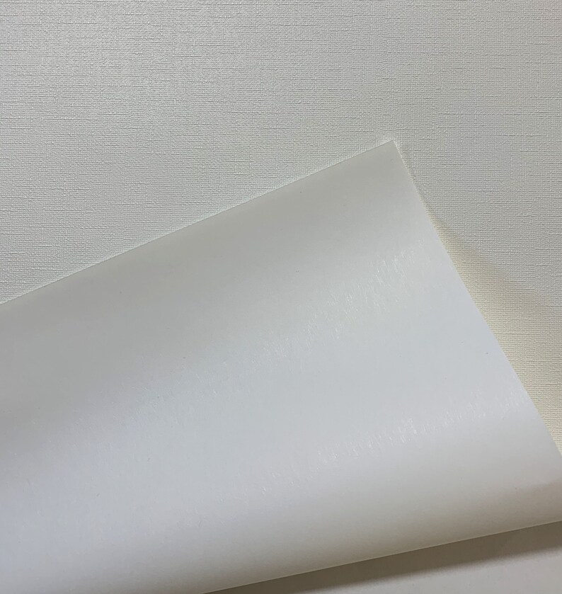 K&ouml;nnte beinhalten: Nahaufnahme von zwei wei&szlig;en Papieroberfl&auml;chen. Eine Oberfl&auml;che hat ein strukturiertes, leinenartiges Aussehen, w&auml;hrend die andere glatt und gl&auml;nzend ist. Das gl&auml;nzende Papier ist teilweise abgezogen und gibt die Textur der anderen Oberfl&auml;che frei. Die Beleuchtung erzeugt Glanzlichter und Schatten.
