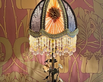 May include: An antique table lamp with a floral design. The lampshade is a dark blue and gold color with crystal bead fringe. The base is bronze with floral accents, sitting on a round wooden table with a decorative doily.