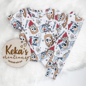 Puede incluir: Conjunto de pijama de punto acanalado blanco con estampado de personajes de dibujos animados con gorros navideños y otros elementos festivos. El conjunto incluye una camiseta de manga corta y un pantalón. También hay un letrero de madera con el texto "Keka's creationsp Handmade with Love".