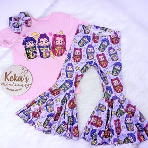 May include: A pink short-sleeved t-shirt with a graphic of three cartoon characters in cups, paired with matching patterned bell-bottom pants and a bow. The pants and bow feature the same character design. A wooden sign is also in the image.