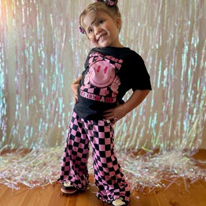 Puede incluir: Un conjunto infantil con una camiseta negra con un gr&aacute;fico de carita sonriente rosa y el texto "THREEENAGER". El conjunto incluye pantalones acampanados a cuadros rosas y negros. La ni&ntilde;a tambi&eacute;n lleva un lazo para el pelo a juego.