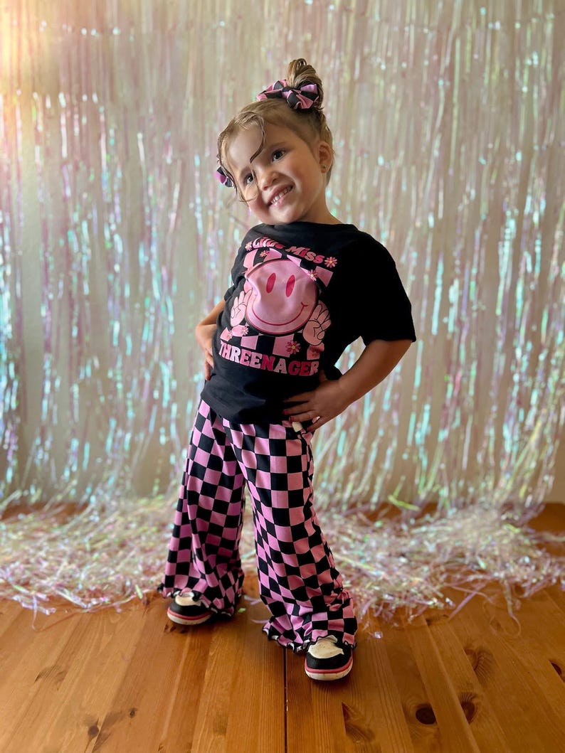 Puede incluir: Un conjunto infantil con una camiseta negra con un gr&aacute;fico de carita sonriente rosa y el texto "THREEENAGER". El conjunto incluye pantalones acampanados a cuadros rosas y negros. La ni&ntilde;a tambi&eacute;n lleva un lazo para el pelo a juego.