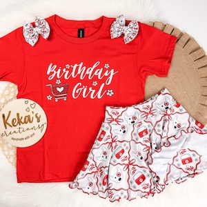 Puede incluir: Una camiseta roja de manga corta con el texto blanco Birthday Girl y un gráfico de carrito de compras, junto a una falda blanca con un patrón repetitivo de caras de perro y carritos en rojo y blanco, además de dos lazos para el cabello.