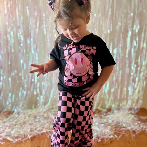Puede incluir: Un conjunto infantil con una camiseta negra con una carita sonriente rosa y el texto "Little Miss Threenager". El conjunto incluye pantalones a cuadros rosas y negros y lazos para el pelo a juego. El conjunto es brillante y alegre.