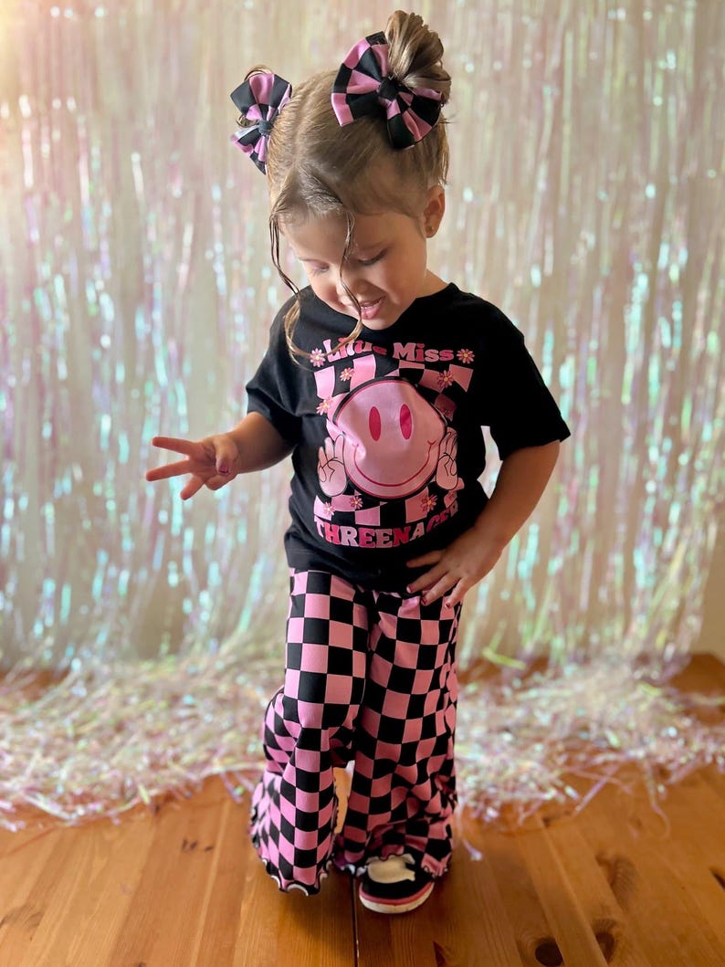 Puede incluir: Un conjunto infantil con una camiseta negra con una carita sonriente rosa y el texto "Little Miss Threenager". El conjunto incluye pantalones a cuadros rosas y negros y lazos para el pelo a juego. El conjunto es brillante y alegre.
