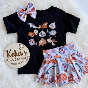 Peut inclure: Ensemble de t-shirt noir, jupe et nœud papillon avec des personnages de poissons de dessins animés. Le t-shirt présente une impression graphique de divers poissons. La jupe et le nœud papillon ont un motif assorti. Un panneau en bois avec le texte "Keka's creations" est également présent.