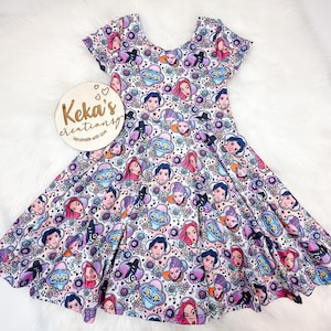 Puede incluir: Vestido de manga corta con un estampado colorido de personajes de dibujos animados, flores y notas musicales sobre un fondo blanco. El vestido tiene un escote redondo y una falda acampanada. La marca "Keka's creations" está en un letrero de madera.