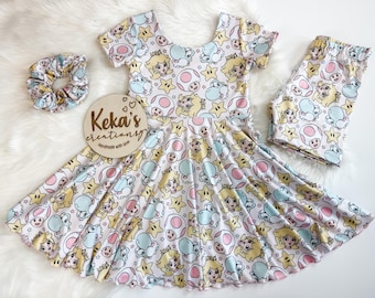 May include: A children's clothing set featuring a dress, shorts, and scrunchie, all adorned with a colorful cartoon character print. The set includes a dress with a flared skirt, matching shorts, and a scrunchie. The wooden sign says "Keka's creations".