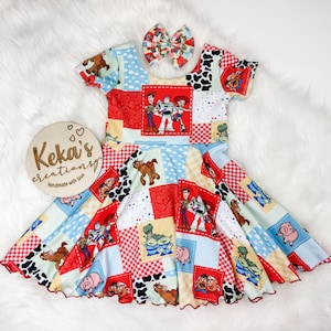 May include: A patchwork dress featuring characters from the Toy Story movie, with a matching bow. The dress has a red, white, blue, and black color scheme, with images of Woody, Buzz Lightyear, and other characters. The dress is designed for children.
