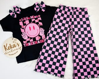 May include: A black t-shirt with a pink smiley face graphic and the text "Little Miss THREENAGE". The outfit includes matching pink and black checkered pants and two hair bows. A wooden sign with the text "Keka's creations" is also included.