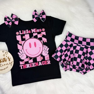 Puede incluir: Un conjunto de camiseta y falda negras con lazos para el pelo a juego. La camiseta presenta una carita sonriente rosa y el texto "Little Miss Threenager". La falda y los lazos tienen un estampado de tablero de ajedrez rosa y negro.