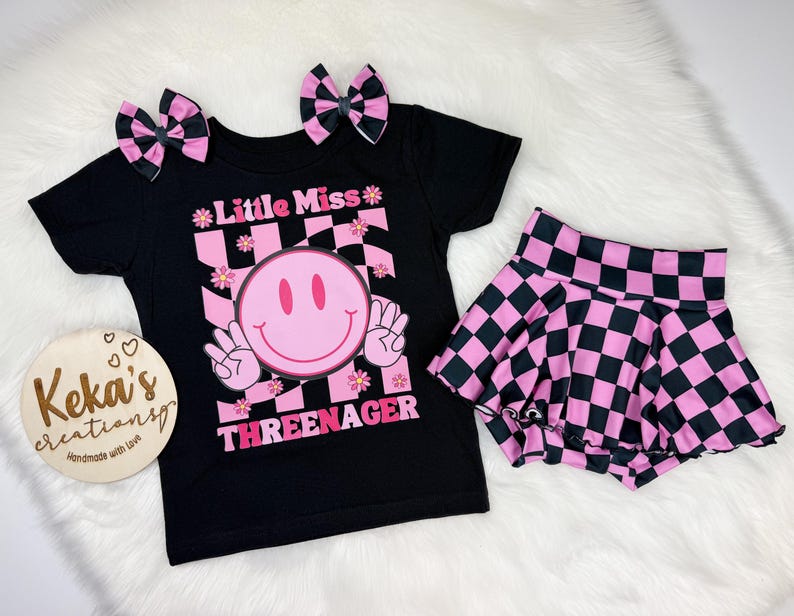 Puede incluir: Un conjunto de camiseta y falda negras con lazos para el pelo a juego. La camiseta presenta una carita sonriente rosa y el texto "Little Miss Threenager". La falda y los lazos tienen un estampado de tablero de ajedrez rosa y negro.