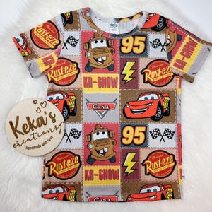 May include: A short-sleeved, patterned t-shirt featuring Cars characters, including Lightning McQueen and Mater, with the text "KA-CHOW" and "Rust-eze". The shirt has a round neckline and is made of a soft, ribbed fabric. A wooden sign with the text "Keka's creations" is in the image.