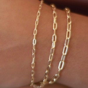Pu&ograve; includere: Tre braccialetti a catena dorati impilati sul polso di una persona. Le catene sono fatte di piccoli anelli ovali.