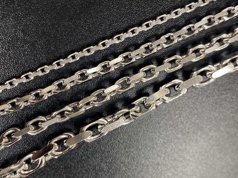May include: Close-up of four silver-colored chain necklaces with different link styles. The chains are arranged diagonally against a dark background, showcasing the variations in design and texture. The chains are made of metal.
