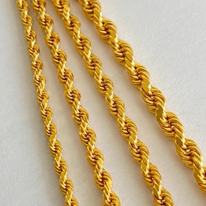 May include: Close-up of four gold-coloured rope chains. The chains are arranged diagonally across the frame, showcasing their twisted design. The chains have a shiny, reflective surface, and the background is a soft, neutral colour.