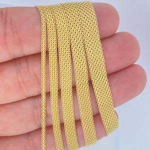 May include: Close-up of several strands of gold-colored mesh chain. The chains are held in a hand against a white background. The mesh pattern is visible, and the chains appear to be made of a shiny, metallic material. The chains are likely used for jewelry making.