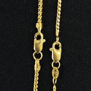 May include: Close-up of two gold-colored chain necklaces with lobster claw clasps. The chains have a delicate, textured design. The background is a solid black color.