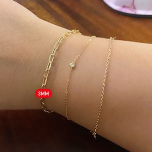 Pu&ograve; includere: Tre braccialetti a catena d'oro sul polso di una persona. Il braccialetto superiore &egrave; una catena a maglie da 3 mm. Il braccialetto centrale ha un piccolo accento di diamante. Il braccialetto inferiore &egrave; una catena delicata.