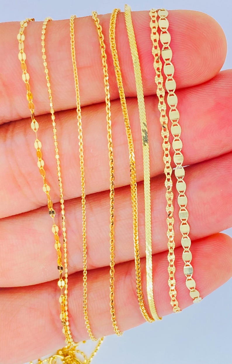 May include: Close-up of several delicate gold necklaces with various chain styles. The chains include oval links, thin strands, and a herringbone design. The jewelry is displayed against a light background.