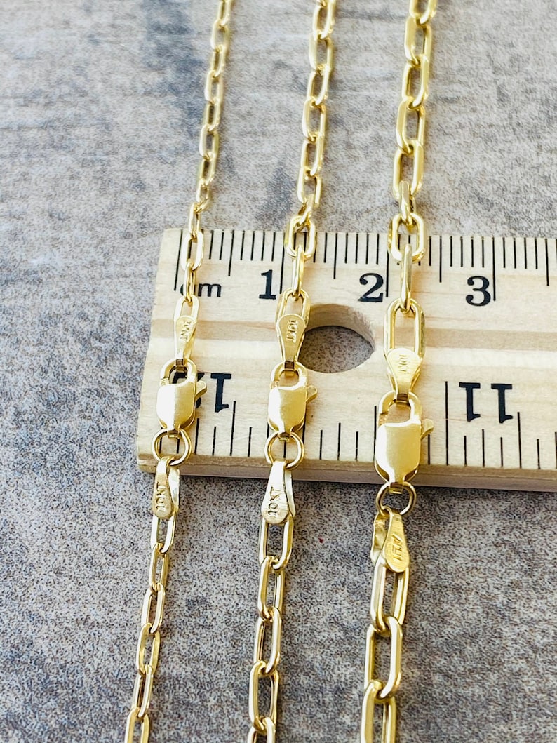 May include: Three gold chains of different lengths, laid out on a wooden ruler. The chains are all the same style, with a flat, oval-shaped link. The ruler is marked in inches and shows that the chains are 18 inches, 20 inches, and 22 inches long.